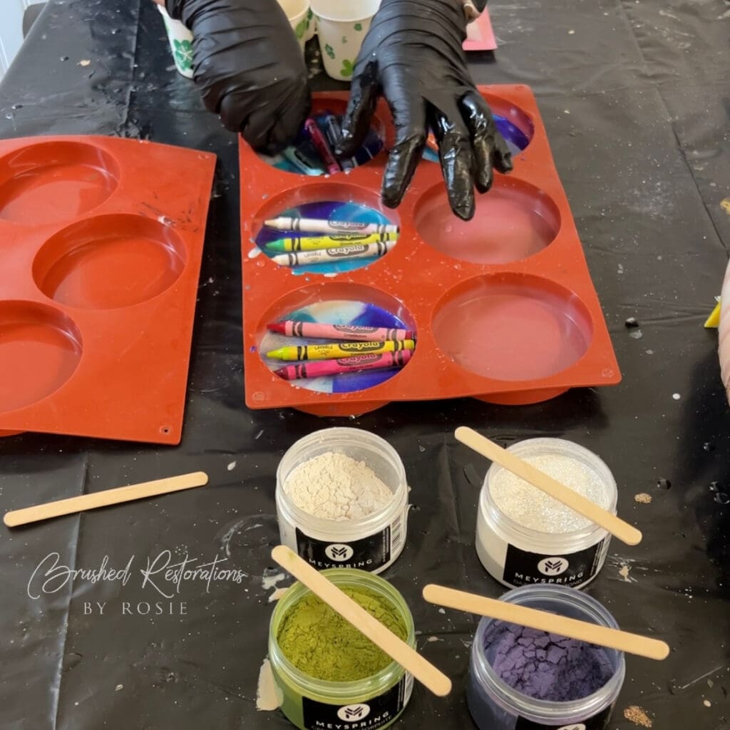 Hands preparing resin materials, including crayons in silicone molds and various colored mica powders. A creative setup for DIY resin crafts, highlighting the initial stages of making resin DIY crayon coasters.