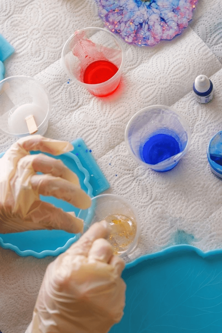 Overhead view of a resin art workspace with various silicone molds, cups of mixed resin in red and blue, bottles of pigment, and completed resin art pieces adorned with gold leaf and glitter.