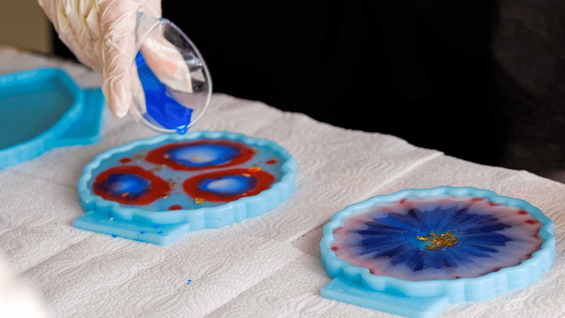 Overhead view of a resin art workspace with various silicone molds, cups of mixed resin in red and blue, bottles of pigment, and completed resin art pieces adorned with gold leaf and glitter.