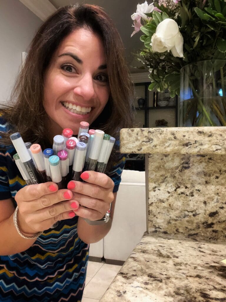 Smiling female artist holding a collection of Copic markers in front of a flower arrangement.