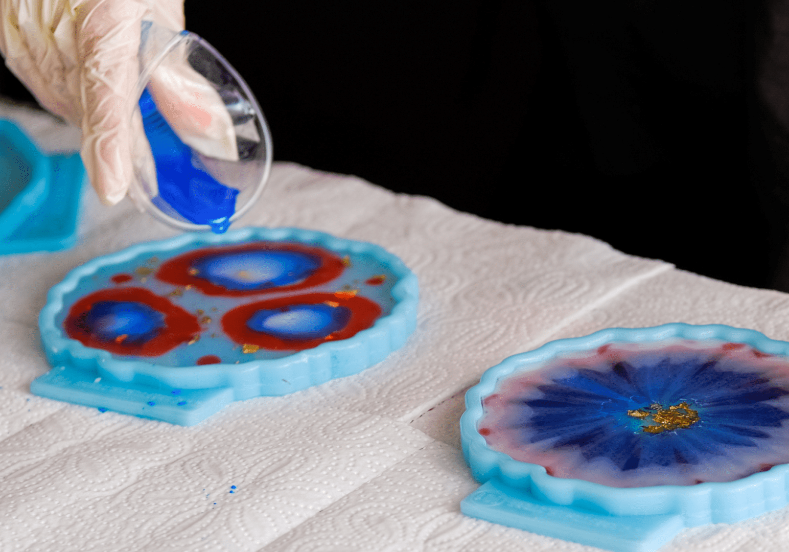 Overhead view of a resin art workspace with various silicone molds, cups of mixed resin in red and blue, bottles of pigment, and completed resin art pieces adorned with gold leaf and glitter.