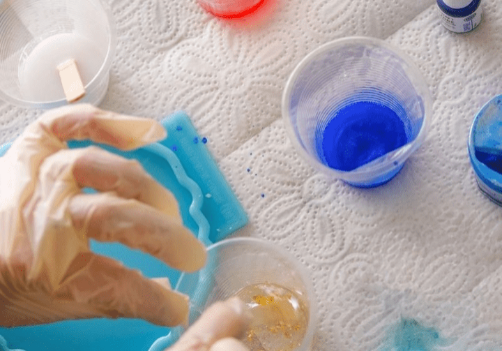 Overhead view of a resin art workspace with various silicone molds, cups of mixed resin in red and blue, bottles of pigment, and completed resin art pieces adorned with gold leaf and glitter.